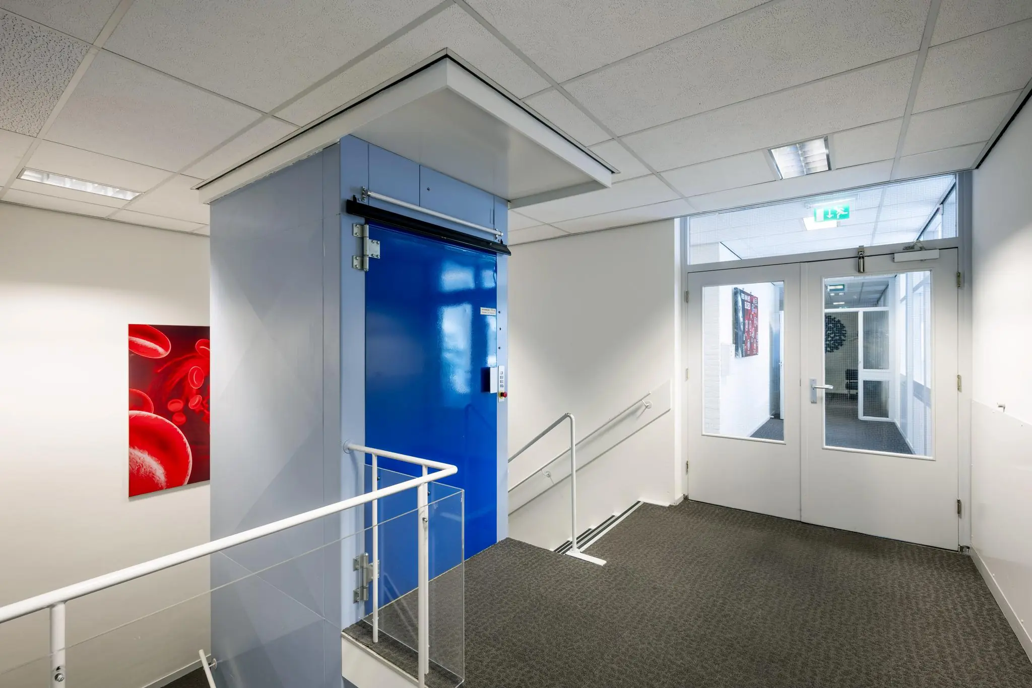 Hal met blauwe interne goederenlift en dubbele glazen deur in een modern kantoorgebouw aan de Jan Tooropstraat.